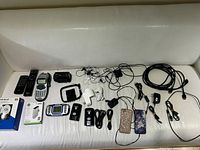 Photo showing various electronics and accessories laid out on a white couch, including remotes, cables, earphones and small electronic devices.