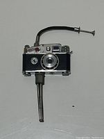 Front view of vintage miniature camera lighter showing lens details, red button, flexible pipe, and nozzle.