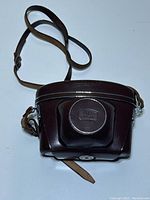 Brown leather camera case closed, showing embossed logo on front and shoulder strap.