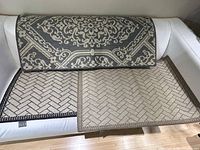 Three woven throw rugs displayed on a white sofa: a dark gray rug with beige floral and geometric pattern, a beige rug and a gray rug each with a herringbone weave pattern.