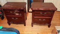 Front view of two wooden bedside tables side by side showing two drawers on each with metal handles and visible surface scratches.
