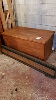 Side angle showing wooden storage chest with a metal baseboard heater in front, both placed on basement floor.