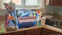 Corner countertop area showing multiple spray bottles with various cleaning solutions arranged behind plastic storage bins and baskets, along with some cleaning brushes and sponges.