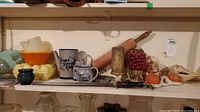 Shelf displaying various kitchen utensils and decorative ceramic items including mugs, rolling pin, candles and figurines