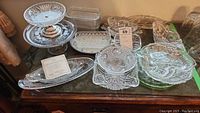 Overview of displayed crystal and glass serving ware on wooden table showing variety of pieces including trays, pedestal bowls, and covered dishes.