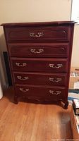 Front view of five drawer highboy dresser showing brass handles and reddish-brown wooden finish with some wear.
