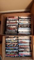 Top view of two boxes filled with assorted DVDs showing movie titles including John Wayne westerns, Frozen, Bridesmaids, and action films like The Expendables and Skyfall.