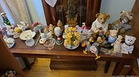 Overall view of various figurines and faux flower arrangements spread over a wooden table, showcasing teddy bear banks, animal figurines, and decorative floral pieces.