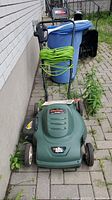Front view of the Black & Decker electric lawn mower with green extension cord wrapped on handle, beside trash bin on outdoor patio.