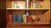 Wide view of two shelves containing various glasses, including fifteen colorful tall MCM style glasses and three clear beer glasses.