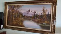 Framed oil painting showing a river flowing through a wooded area towards snowy mountains under a pale sky, with a cabin on the riverbank.