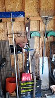 Photo of garden tools hung on wooden wall including rakes, snow rake, shovel, saw, with some tools leaning against the wall.