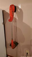 Full view of Black and Decker cordless trimmer hanging on wall, showing orange body, black guard, and metal shaft.