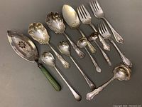 Photo of entire set of 10 antique silver plated serving utensils showing various types including pierced pie/cake server with green handle, serving spoons, forks, and ladles all with ornate floral designs.