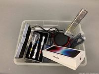 Photo showing the lot items inside a plastic bin including the tubular lock tools in metal case, microphone, wireless speaker, and iPhone box.