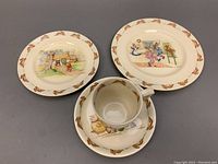 Set of four Bunnykin themed ceramic pieces including three plates and a cup with saucer, showing bunny designs and borders.