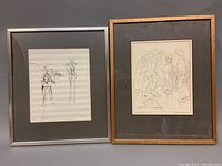 Two framed sepia pen & ink sketches side by side on a gray surface with one frame's glass cracked.