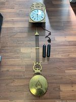 Photo showing disassembled clock components including the enamel dial with Roman numerals, gilded bas-relief clock housing top, brass pendulum with lyre design, metal chains, and two black clock weights on wood floor.