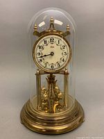 Full view of the clock with glass dome on. Clock is brass with a round white face with black numerals and floral designs, four brass pillars, and four rotating balls beneath the face. Base is brass and circular.