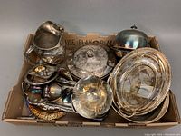 Full box view of all silver plate servingware items in a cardboard box, showing pitcher, covered butter dish, ashtray, trays and bowls.