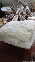 Photo of folded queen size white duvet alongside visible pillow shams and bedspread in background, all showing some stains.