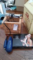 Blue hair dryer, silver curling iron/flat iron, and Forever Flawless manicure kit box with image of a woman, displayed on floor beside a basket and large cardboard box.