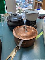 Photo showing copper pots, black ceramic bean pot with lid, and white enamel cast iron fondue set on table with background view