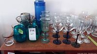 Wide shot of assorted glasses including wine glasses with dark stems, greenish goblet-style glasses, teal colored glasses, tall blue pitcher and candle holder with candle on a wooden surface