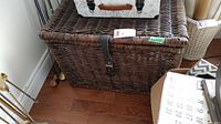 Wicker chest showing front with leather strap and metal clasps, placed on wooden floor near corner of wall.