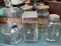 Set of six glass jars on countertop, some with brass and silver lids, including round and square shapes
