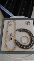 Overview of various jewellery pieces arranged in a black display tray including necklaces, bracelets and earrings in silver, gold-tone, black and blue bead accents.