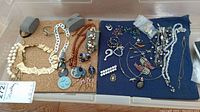 Full view of assorted costume and fine jewelry including necklaces, bracelets, brooches, earrings, gold chains, and enamel pins on cork and fabric trays.