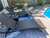 Two identical dark brown synthetic wicker outdoor loungers placed side by side on a patio deck next to a pool, showing entire loungers and part of the outdoor setting.