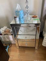 View of the full night stand with three drawers and reflective beveled mirrored surfaces, items on top include a clear glass lamp and other miscellaneous objects.