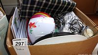 Box of patio dishware including plastic bowls, enamelled plates, mugs, flatware, vinyl tablecloth, and candle holders.