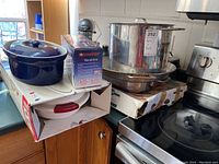 Overview of lot on counter showing stainless steel pot on stove, roasting and paella pans, boxed StarFrit Mandoline, Thermos server, and blue ceramic casserole dish on boxed Thermos server.