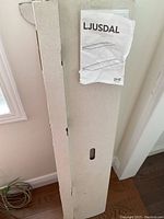 IKEA LJUSDAL wall shelf packed flat in its original cardboard packaging leaning against the wall. Instruction manual labeled LJUSDAL resting on top.
