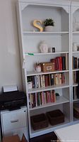 Full view of white wooden shelf with six shelves containing decorative items and books. Shelf is tall with scalloped top edges.