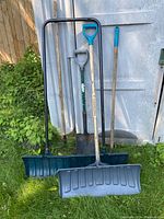Full view of the five garden and snow tools showing two rakes, one shovel, and two snow shovels against a metal door outdoors.