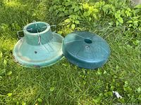 Green metal Christmas tree stand and green plastic patio umbrella stand placed on grass with foliage backdrop showing condition and details.