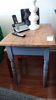 Front view of pine end table showing natural wood top with some scratches, blue-gray painted legs and base, with cordless phone and lamp on top.