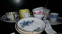 Front view showing various vintage floral teacups on their matching saucers with patterns mainly blue, yellow, or multicolor flowers with gold trim