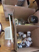 Three moving boxes filled with various kitchen and dining items including glass, ceramic, and wooden pieces. Visible white plastic dish drainer and several glass bottles with metal lids.