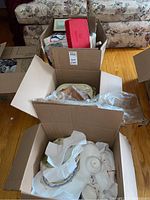 Three boxes containing various vintage kitchen items including dishes, wood rolling pin, plungers, trays, bowls, cups, and saucers.