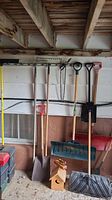 Full assortment of garden tools hanging on a wall including shovels, rakes, and trimmers, plus a woodpecker birdhouse on the floor