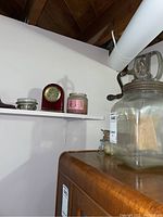 Shelf view showing small red clock, pink ribbon tin candle, small glass jar and butter churn on wooden surface