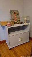 Grey media stand with cupboard and shelf, displaying a framed needlepoint of a girl, box of costume jewelry, two scented candles, a glass preserving jar, and a decorative glass bowl on top.
