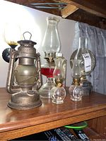 Overview of five oil lanterns on wooden surface showing variation in size, material and design. Includes two smaller matching glass lanterns, larger glass lanterns with oil and metal lantern.