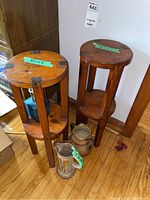 Two wooden plant stands with round tops and lower shelves, one stand has label showing dimensions 8 x 23 inches. Two pottery jugs with handles placed in front of stands, showing some wear.