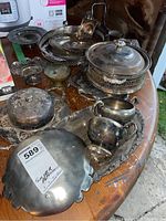 Various silver plated serving items spread on a wooden table, including trays, butter dish cover, creamer and sugar, small dishes and serving pieces, all showing signs of wear and tarnish.